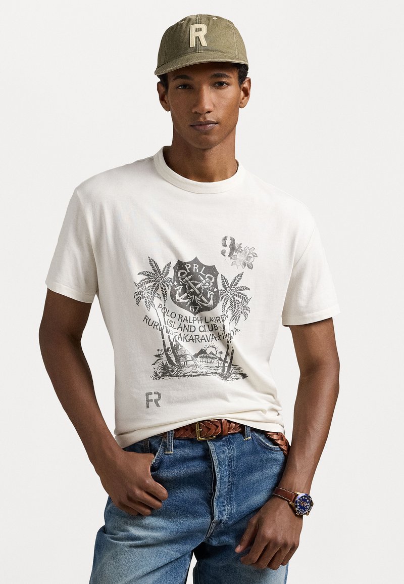 Balta kokvilnas T-kreksa ar melnu grafisku attēlu, kurā redz palmās un enkurus. Apvienots ar gaiši ziliem džinsiem un brūnu austu jostu.