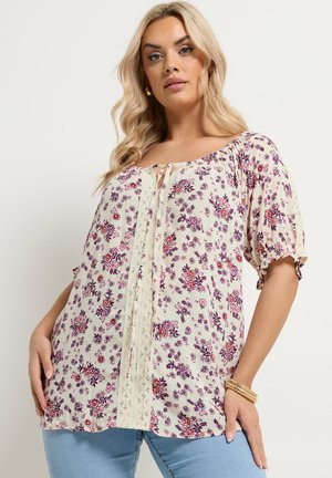 Donna bionda che indossa una blusa color crema con stampa floreale viola e rossa, maniche a sbuffo, dettaglio in pizzo sul davanti e jeans azzurri, posando davanti a un muro bianco.