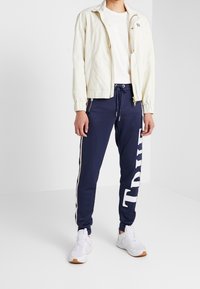 Ljust beige zip-jacka över en vit t-shirt, kombinerad med marinblå joggers med vit "TRUE" text och sidostreifen, bärs med vita sneakers.