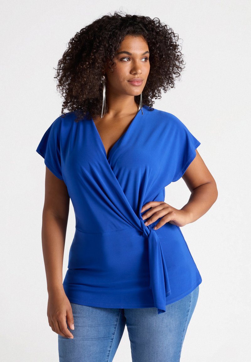 Top blu in tessuto liscio con scollatura a V profonda, maniche corte e un nastro annodato in vita, abbinato a jeans chiari.