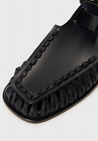 Sandal nera con una texture intrecciata sulla fascetta del piede e una tomaia in pelle liscia. Punta arrotondata con una suola piatta e resistente.