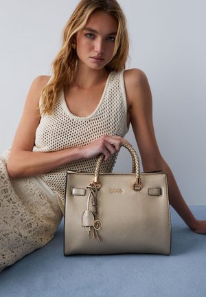 Mlada ženska v bež kvačkanem vrhu in krilu drži teksturirano zlato torbico z vtkanimi ročaji in obeskom na modri površini.