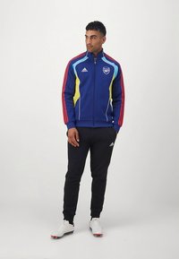 Blauwe sportjas met rode strepen en gele accenten, ritssluiting, logo op de borst; gecombineerd met zwarte broek en witte sportschoenen.