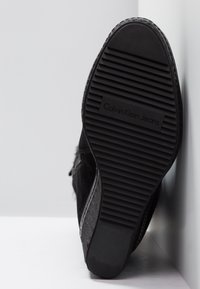 Semelle d'une chaussure noire Calvin Klein Jeans avec des rainures horizontales et un talon texturé, reposant contre une surface blanche.