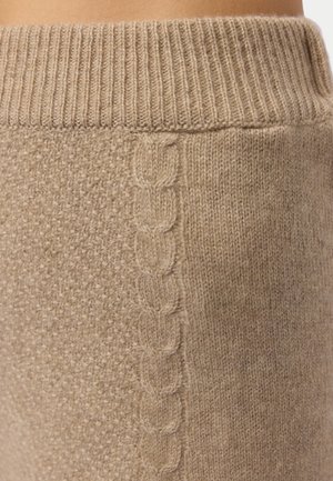 Nahaufnahme eines beige gestrickten Stoffs mit geripptem Bund, strukturiertem Muster auf der linken Seite und vertikalem geflochtenem Zopfmuster in der Mitte.