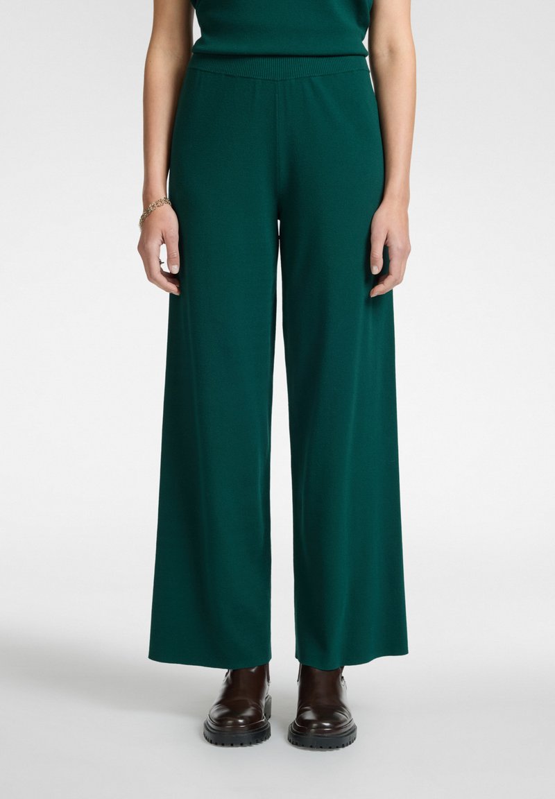 Pantalones de pierna ancha verde oscuro hechos de una tela suave con una cintura elástica, combinados con botas de tobillo marrones con cordones.