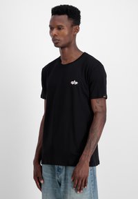 Sort bomulds T-shirt med rund halsudskæring, der har et lille broderet stjernesymbol i hvidt og rødt på brystet. Korte ærmer.