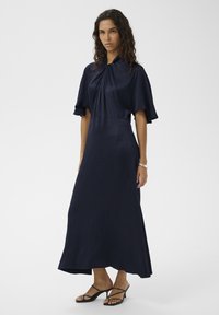 Mornarsko modra maksi obleka s kratkimi razširjenimi rokavi, nabranim pasom, teksturirano tkanino in ravnim silhueto. Nošena s črnimi sandali z naramnico.