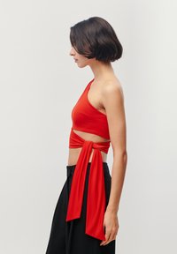 Top corto rosso monospalla con dettaglio legato in vita, realizzato in tessuto morbido. Il design presenta una sezione con taglio laterale.