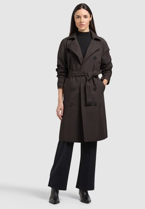 Naine, kes kannab tumepruuni vöökinnitusega trençkot, musta kõrge kaelusega pluusi, tumedaid laiade sääreosadega pükse ja musti pahkluu saapaid, seisab halli tausta ees.