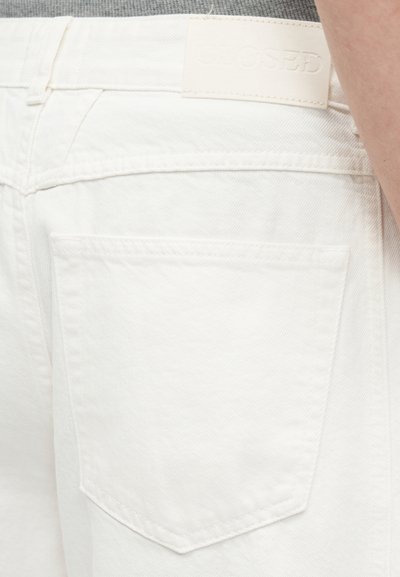 Gros plan sur un jean en denim blanc montrant la poche arrière, la passoire de ceinture, et une vue partielle du bras d'une personne sur le côté.