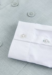 Vit skjortärm med märkesmanschettknappar som vilar på ljusgrått texturerat tyg med grå knappar.