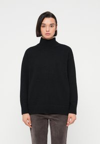 Femme aux cheveux courts et foncés portant un pull à col roulé noir côtelé et un pantalon gris foncé contre un fond blanc uni.