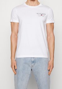 Bílý bavlněný tričko s kulatým výstřihem a krátkými rukávy. Na hrudi se nachází černě tištěné logo "HILFIGER EST. 1985 NEW YORK".