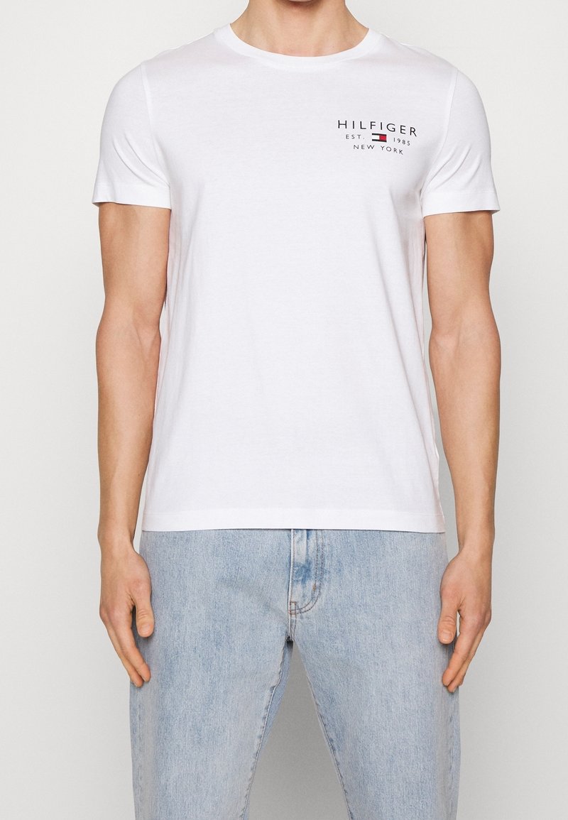 Bílý bavlněný tričko s kulatým výstřihem a krátkými rukávy. Na hrudi se nachází černě tištěné logo "HILFIGER EST. 1985 NEW YORK".