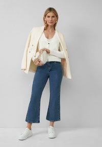 Witte button-up blazer over een crèmekleurige V-hals top, gecombineerd met donkerblauwe cropped jeans en witte sneakers. Houdt een ronde clutch bij zich.