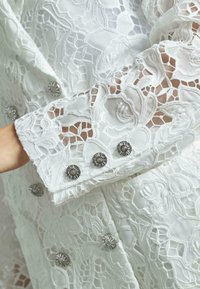 Giacca bianca in pizzo con intricati motivi floreali, tessuto testurizzato e bottoni a forma di fiore argentati lungo i polsini.