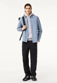 Camisa de cuadros azul y blanca, sudadera gris debajo, pantalones cargo negros y zapatillas negras. El modelo lleva una mochila sobre un hombro.