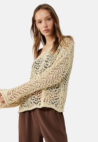 Maglione a maglia aperta di colore crema, con maniche lunghe e una vestibilità ampia. Abbinato a pantaloni marroni fluidi; dettagli in filato testurizzato presenti in tutto il capo.