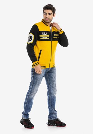 Gelbe und schwarze Zip-Jacke mit gestreiften Akzenten, Logo-Patches und zwei Seitentaschen. Getragen mit blauen Jeans und schwarzen Sportschuhen.
