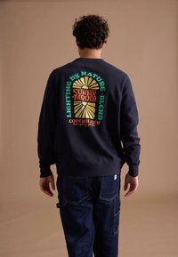 Marineblauwe sweatshirt met kleurrijke print op de achterkant met tekst en afbeeldingen, ronde hals, geribbelde boorden en zoom; gecombineerd met donkere spijkerbroek.
