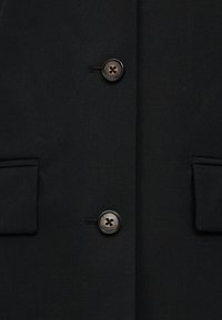 Μαύρο παλτό με δύο σκοτεινά κουμπιά κουμπωμένα και δύο τσέπες με καπάκι στην κάθε πλευρά στο ύψος της μέσης.