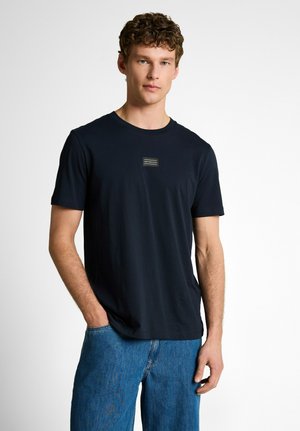 Junger Mann mit lockigem Haar, der ein schlichtes schwarzes T-Shirt und blaue Jeans trägt und mit einer Hand in der Tasche vor einem weißen Hintergrund steht.