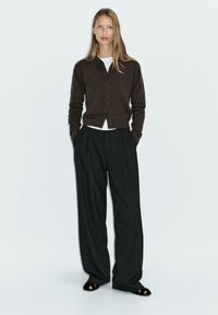 Cardigan marrone con bottoni, top bianco sotto, abbinato a pantaloni a vita alta a righe sottili di colore scuro e sandali neri. Design semplice e aderente.