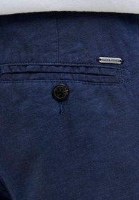 Navyblauwe stof met een gladde textuur, voorzien van een zwarte knoop en een metalen label met de opdruk "JACK & JONES" op de zak.