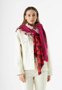 Écharpe géométrique rouge et rose en matériau doux, drapée autour du cou. Portée sur une veste en jean blanche et un pantalon assorti.