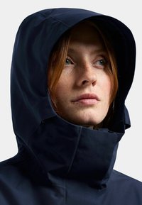 Marinblå vattentät jacka med hög krage och justerbar huva, med en slät, matta tyg och minimalistiska designdetaljer.