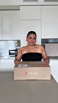 Pessoa com brincos e tatuagem está atrás de um balcão de cozinha segurando uma caixa de entrega da Zalando numa cozinha moderna e branca.