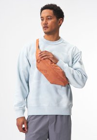 Orangene Nylontasche mit geschwungener Form, zwei Knöpfen, über einer hellblauen Sweatshirt und grauen Hosen quer über die Brust getragen.