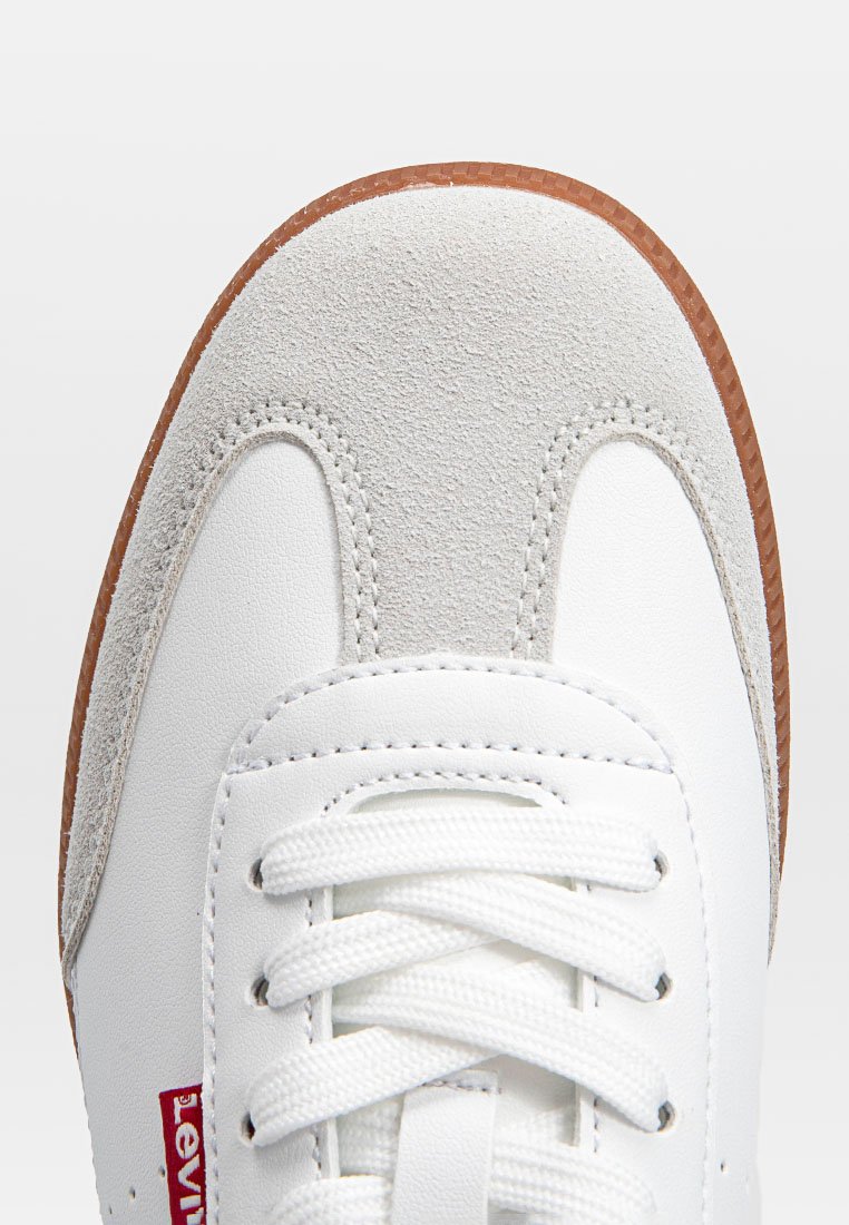 Zapatilla blanca con acentos de ante gris texturizado y suela de goma marrón. Cuenta con cordones planos y material de cuero, mostrando detalles de costura.