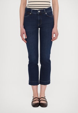 Femme portant un jean court bleu foncé, des ballerines noires à double bride et un top sans manches à rayures horizontales noires et blanches.