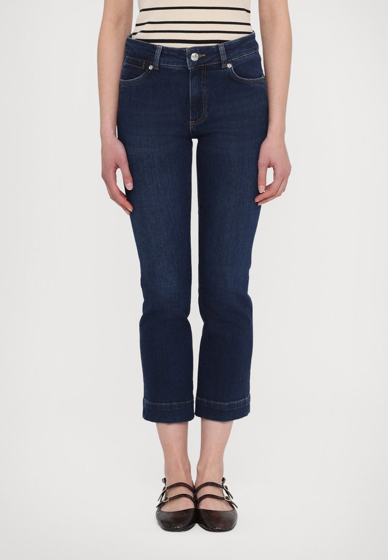Femme portant un jean court bleu foncé, des ballerines noires à double bride et un top sans manches à rayures horizontales noires et blanches.