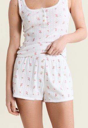 Ensemble de pyjama composé d'un débardeur côtelé blanc et d'un short, ornés de petits motifs de roses roses, avec des boutons sur le haut et le short, porté par une personne.