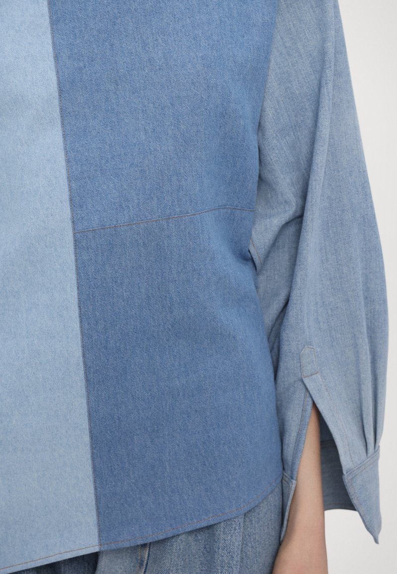 Denim shirt featuring varied shades of blue, a slight texture difference, wide sleeves, and side slits for design detail.