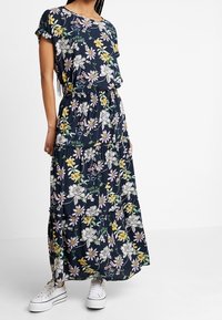 Femme portant une robe maxi à manches courtes à motifs floraux bleu marine, associée à des baskets blanches à plateforme, se tenant devant un fond blanc uni.