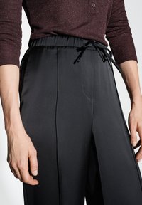 Pantaloni neri a gamba larga con una texture liscia, dotati di vita con cordoncino e davanti plissettato, parzialmente visibili con un top in maglia scuro.