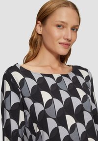 T-shirt à manches longues avec un col rond, présentant un motif géométrique dans des nuances de noir, gris et blanc. Tissu doux avec une texture lisse.