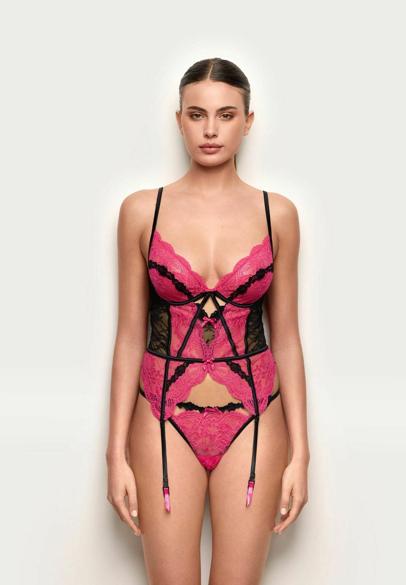 Vrouw staat tegen een lichte achtergrond, draagt roze en zwart kanten lingerie met jarretels, kijkt recht vooruit met een neutrale uitdrukking.