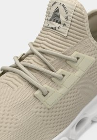 Bézs kötött sneaker texturált felsőrésszel, bézs fűzőkkel, melyek három műanyag szemnyíláson futnak át, valamint fehér talppal kör alakú kivágásokkal.