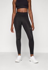 Leggings noirs taille haute en tissu extensible avec des détails de couture et un petit logo blanc sur la jambe inférieure, accompagnés de chaussures de sport blanches.