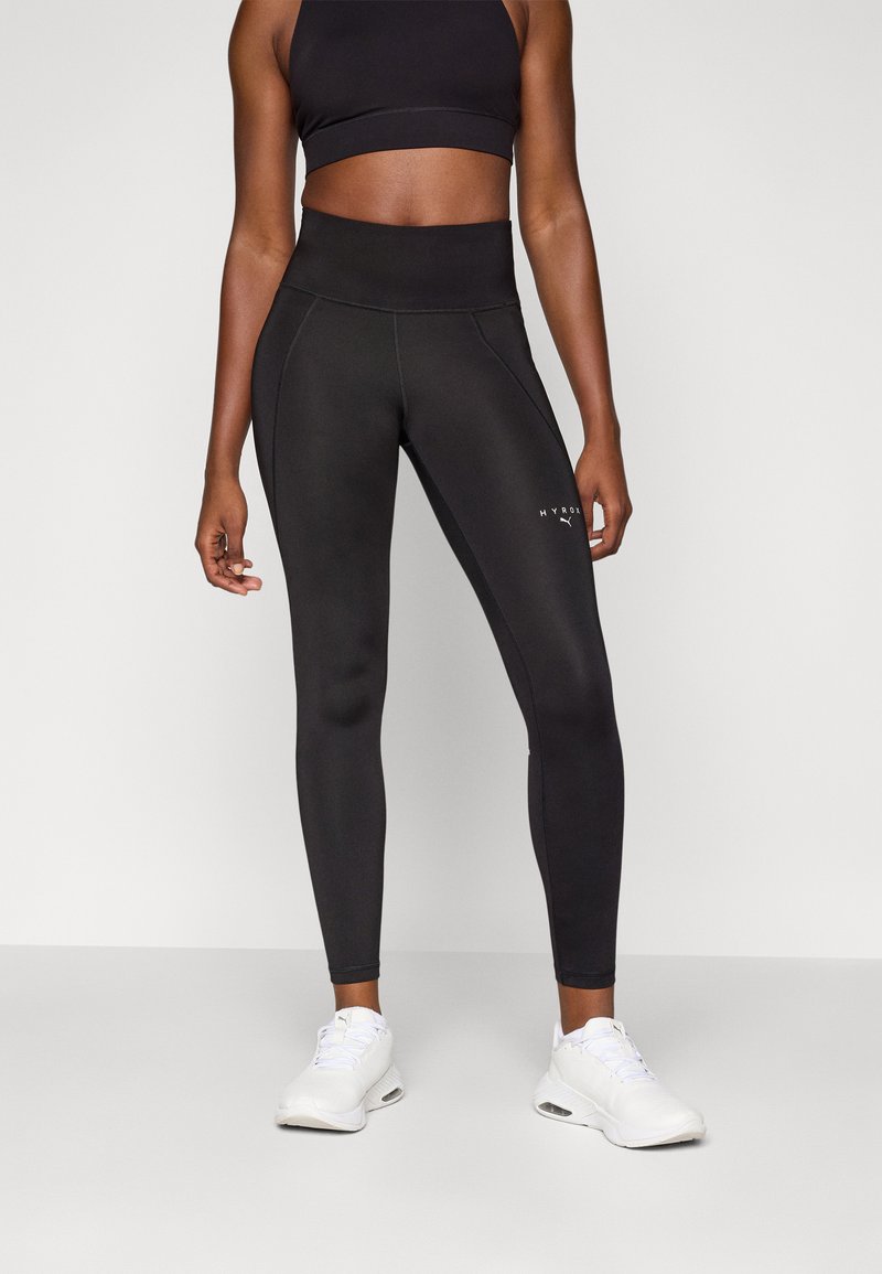 Leggings noirs taille haute en tissu extensible avec des détails de couture et un petit logo blanc sur la jambe inférieure, accompagnés de chaussures de sport blanches.