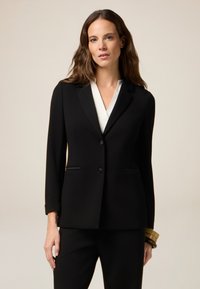 Blazer nero su misura con un solo bottone, rever a punta e tasche a filetto. Indossato sopra una camicetta bianca, abbinato a pantaloni neri. Tessuto liscio.