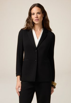 Zwarte op maat gemaakte blazer met een enkele knop, notch revers en weltzakken. Draagt over een witte blouse, gecombineerd met zwarte broek. Soepele textuur.