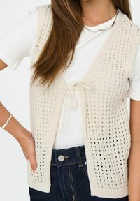 Light beige knitted vest with open weave, featuring a tie front closure and two front pockets. Worn over a white fitted t-shirt.