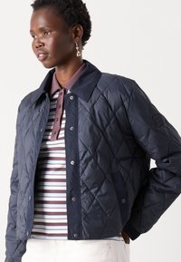 Donkerblauwe gewatteerde jas met drukknoopsluiting en een grote kraag, gedragen over een gestreept poloshirt in bruin- en wittonen.