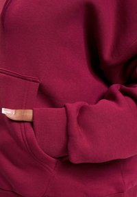 Bordeauxrotes Sweatshirt mit geripptem Bündchen und Tasche. Hergestellt aus weichem Stoff mit glatter Textur, der detaillierte Nähte entlang der Nähte hervorhebt.